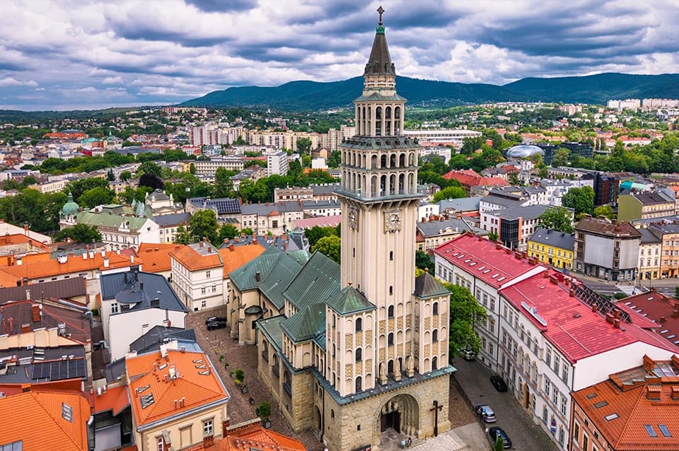 Panorama centrum Bielska-Białej z lotu ptaka. Na pierwszym planie widoczna jest katedra św. Mikołaja z charakterystyczną wysoką wieżą, otoczona zabudową starego miasta. W tle rozciągają się zielone wzgórza Beskidów i zabudowa miejska.