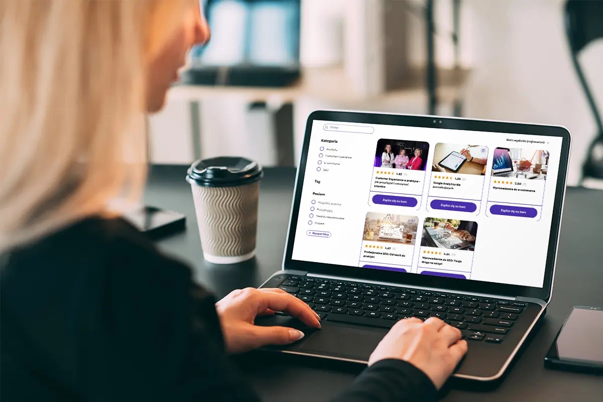 Kobieta siedzi przy biurku i korzysta z laptopa, przeglądając platformę z kursami online; obok stoi kubek kawy i leży smartfon.