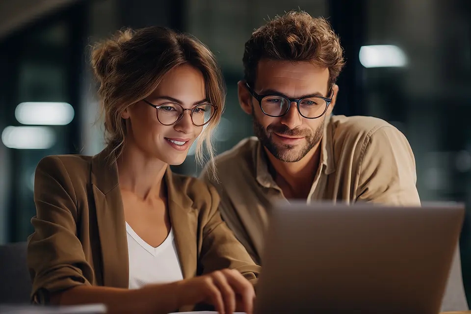 Dwoje młodych dorosłych w okularach, kobieta i mężczyzna, siedzą obok siebie przy laptopie, skupieni na ekranie, w nowoczesnym wnętrzu biurowym wieczorem.