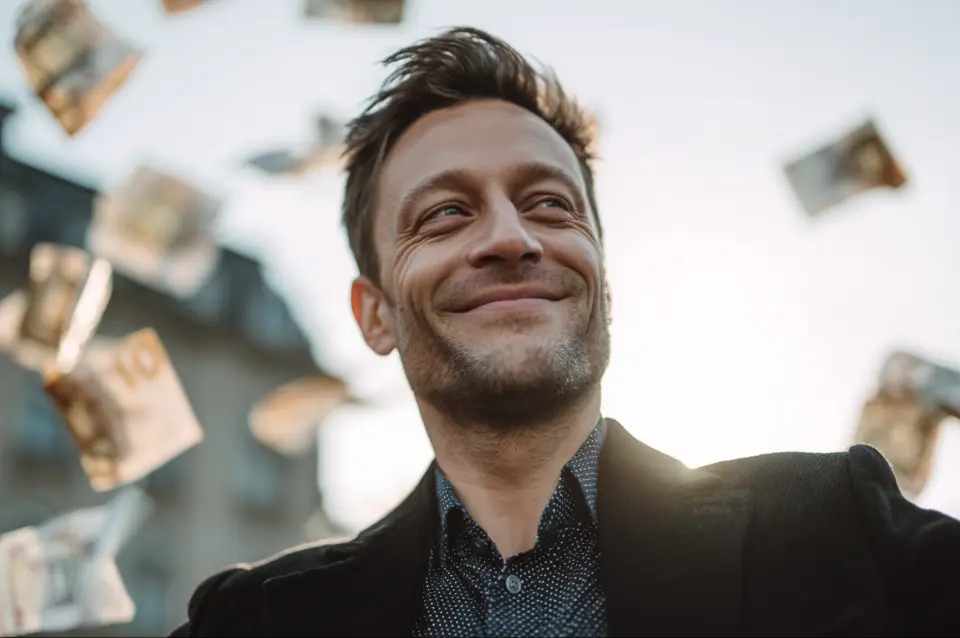 Uśmiechnięty mężczyzna w garniturze stoi na zewnątrz, a wokół niego unoszą się banknoty euro w powietrzu