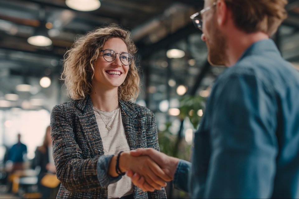 Uśmiechnięta kobieta w okularach podaje rękę mężczyźnie w biurze, witając się w przyjaznej atmosferze