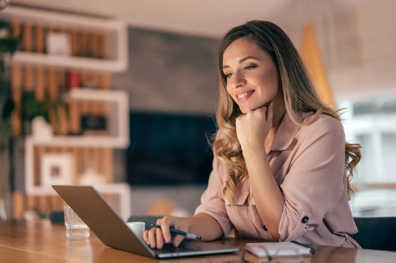 Kobieta siedząca przy biurku i pracująca na laptopie, uśmiechnięta, w domowym biurze