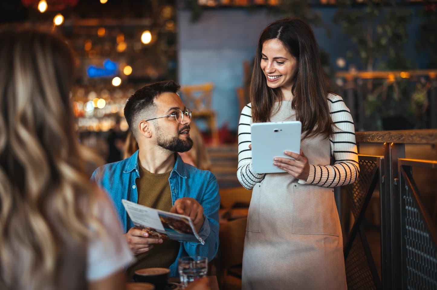 Obsługa restauracji przyjmuje zamówienie za pomocą tabletu od gościa siedzącego przy stoliku. Przytulne wnętrze lokalu z nastrojowym oświetleniem i gośćmi w tle.