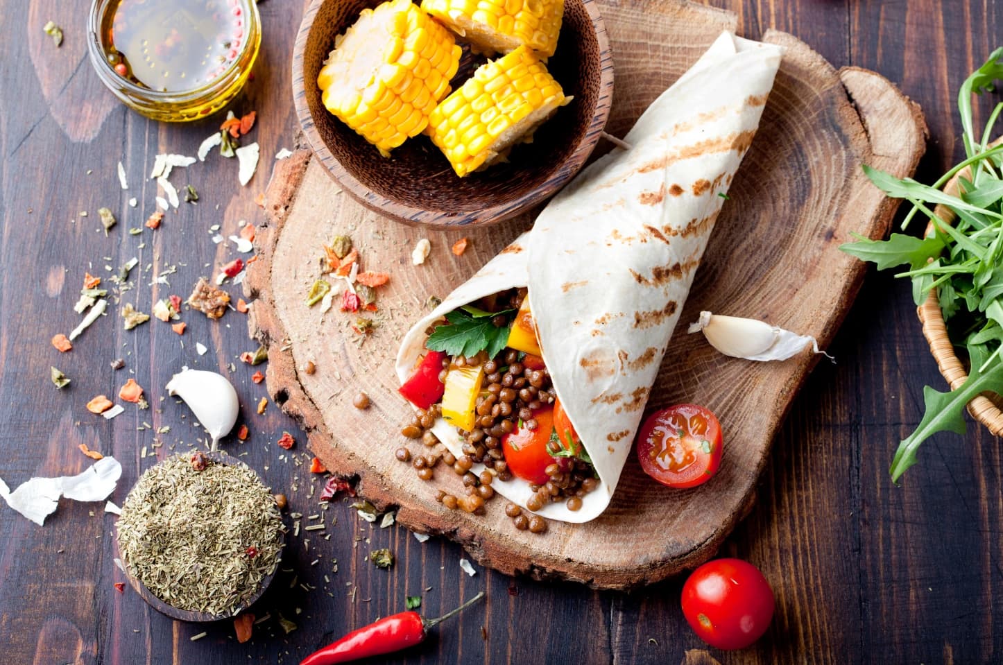 Tortilla wypełniona soczewicą, pomidorkami koktajlowymi, kolorową papryką i świeżymi ziołami, podana na drewnianej desce. Obok znajdują się kawałki kukurydzy, rukola, czosnek, przyprawy oraz miseczka z oliwą na ciemnym drewnianym blacie.