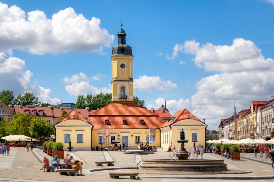 Historyczny budynek z wieżą zegarową przy rynku miejskim, z fontanną na pierwszym planie oraz zabudową i ogródkami po bokach, w słoneczny dzień z błękitnym niebem i chmurami.