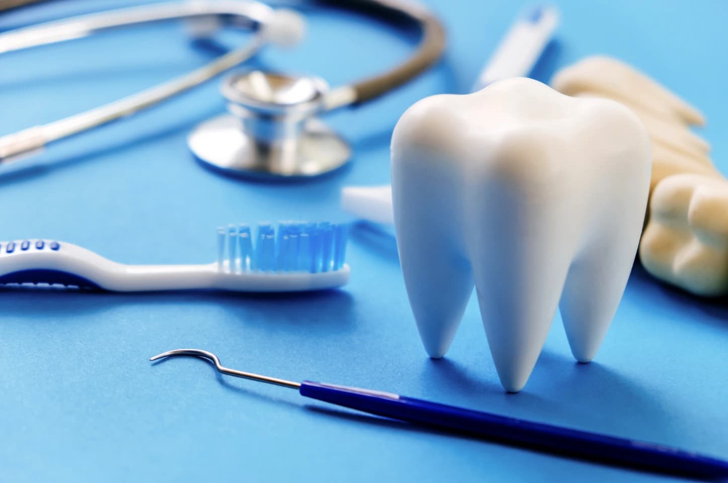 Model zęba ustawiony na niebieskiej powierzchni obok szczoteczki do zębów, lusterka dentystycznego i stetoskopu; w tle widoczne rękawiczki medyczne i narzędzia stomatologiczne.