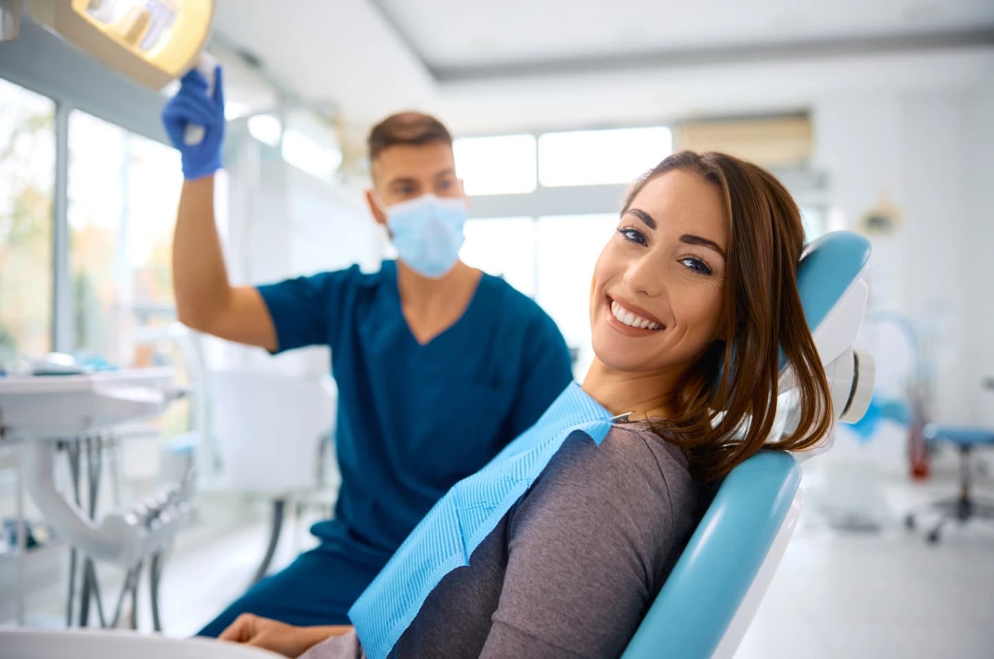 Osoba siedząca na fotelu dentystycznym w gabinecie stomatologicznym, zwrócona w stronę aparatu, z widocznym uzębieniem; w tle inna osoba w stroju medycznym trzymająca lampę zabiegową.