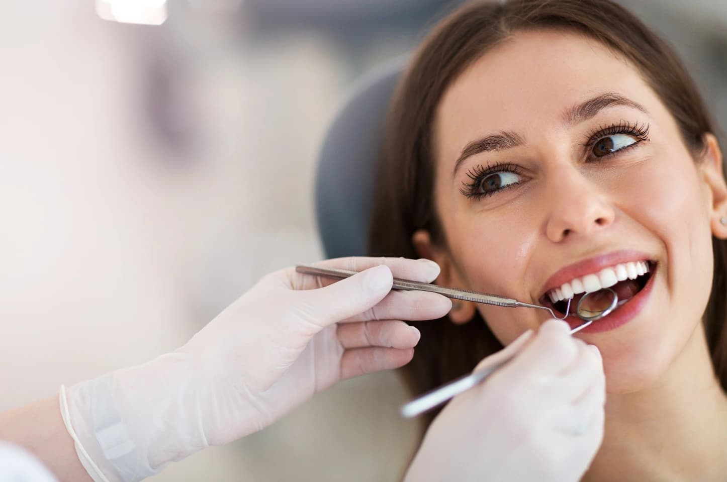 Zbliżenie na osobę siedzącą na fotelu dentystycznym podczas badania stomatologicznego; dłonie w rękawiczkach używają lusterka i narzędzia do kontroli zębów w otwartych ustach, w jasnym gabinecie. 
