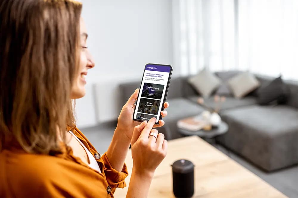 Uśmiechnięta kobieta trzyma smartfon i steruje aplikacją do obsługi inteligentnego głośnika stojącego na stole w salonie.
