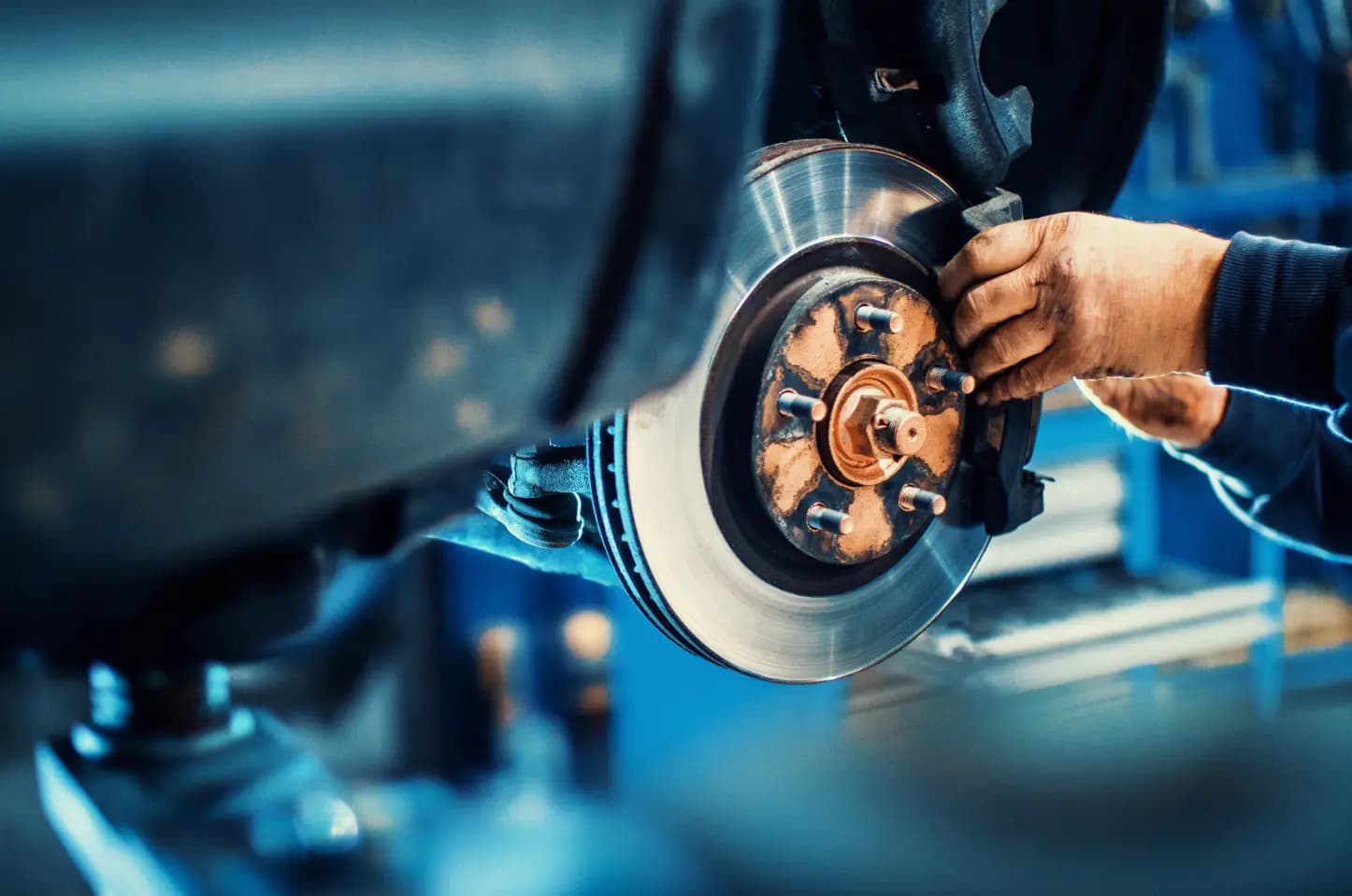 Zbliżenie na tarczę hamulcową i zacisk w uniesionym samochodzie, podczas gdy ręka mechanika pracuje przy układzie hamulcowym w warsztacie.