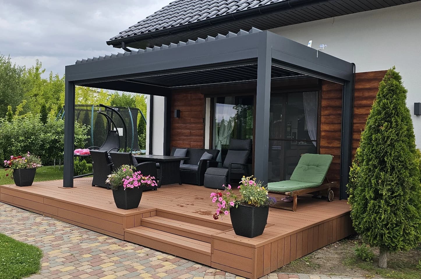 Nowoczesna aluminiowa pergola tarasowa z meblami wypoczynkowymi, zamontowana przy domu na podwyższonym tarasie.